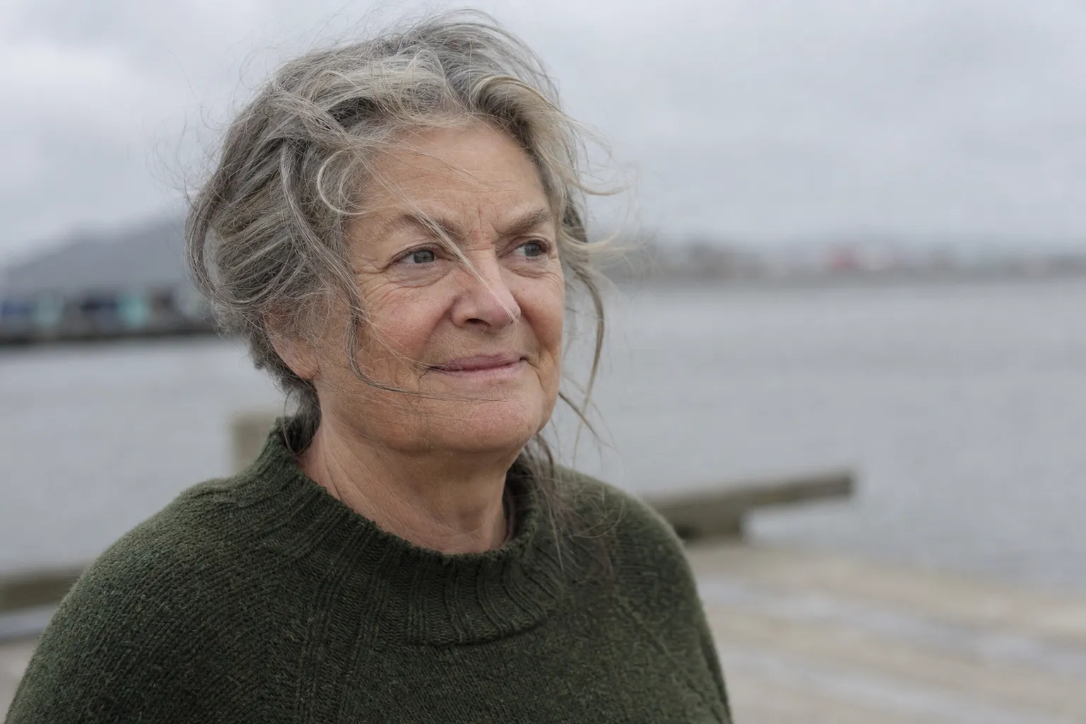 An older woman with windblown grey hair on a wharf, looking out over the water